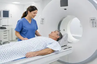 Patient in hospital gown on a CT scanner table, assisted by a radiology technician in a modern, well-lit radiology room.
